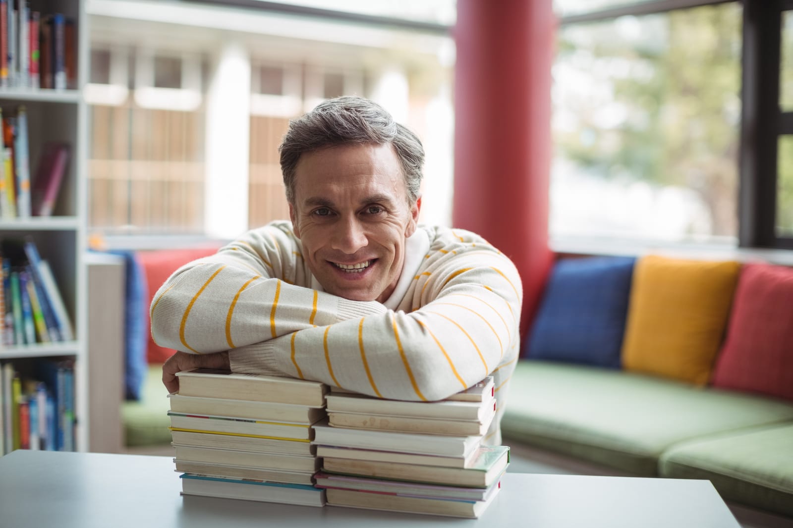 Leitor sorridente apoiado sobre uma pilha de livros em uma biblioteca colorida.