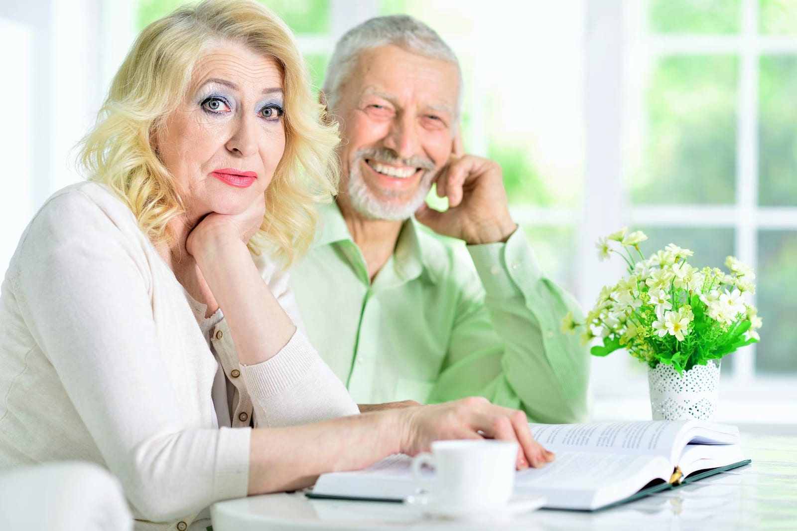 Casal sênior sorridente, folheando um livro juntos à mesa, com uma xícara de café ao lado.